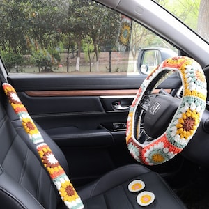 May include: A crocheted steering wheel cover with a colorful floral pattern in shades of orange, yellow, green, white, and brown. The cover is made of yarn and has a granny square design. The steering wheel cover is shown in a car interior with a matching crocheted seatbelt cover and two small crocheted coasters.
