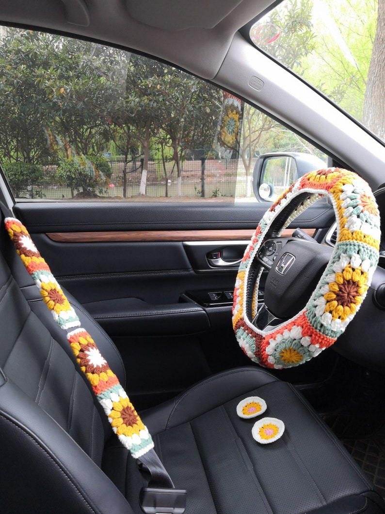 May include: A crocheted steering wheel cover with a colorful floral pattern in shades of orange, yellow, green, white, and brown. The cover is made of yarn and has a granny square design. The steering wheel cover is shown in a car interior with a matching crocheted seatbelt cover and two small crocheted coasters.