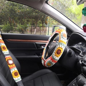 May include: A crocheted steering wheel cover with a floral pattern in shades of yellow, orange, white, and green. The cover is installed on a black leather steering wheel in a car interior.