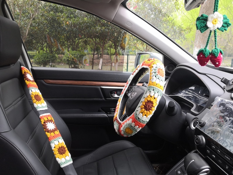 May include: A crocheted steering wheel cover with a floral pattern in shades of yellow, orange, white, and green. The cover is installed on a black leather steering wheel in a car interior.