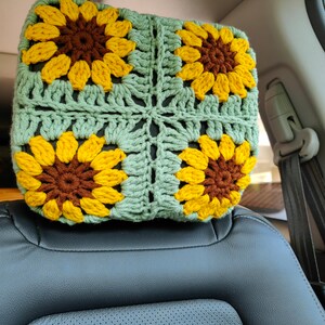 Puede incluir: Un reposacabezas de crochet con fondo verde y cuatro girasoles amarillos con centros marrones. El reposacabezas est&aacute; en un coche.