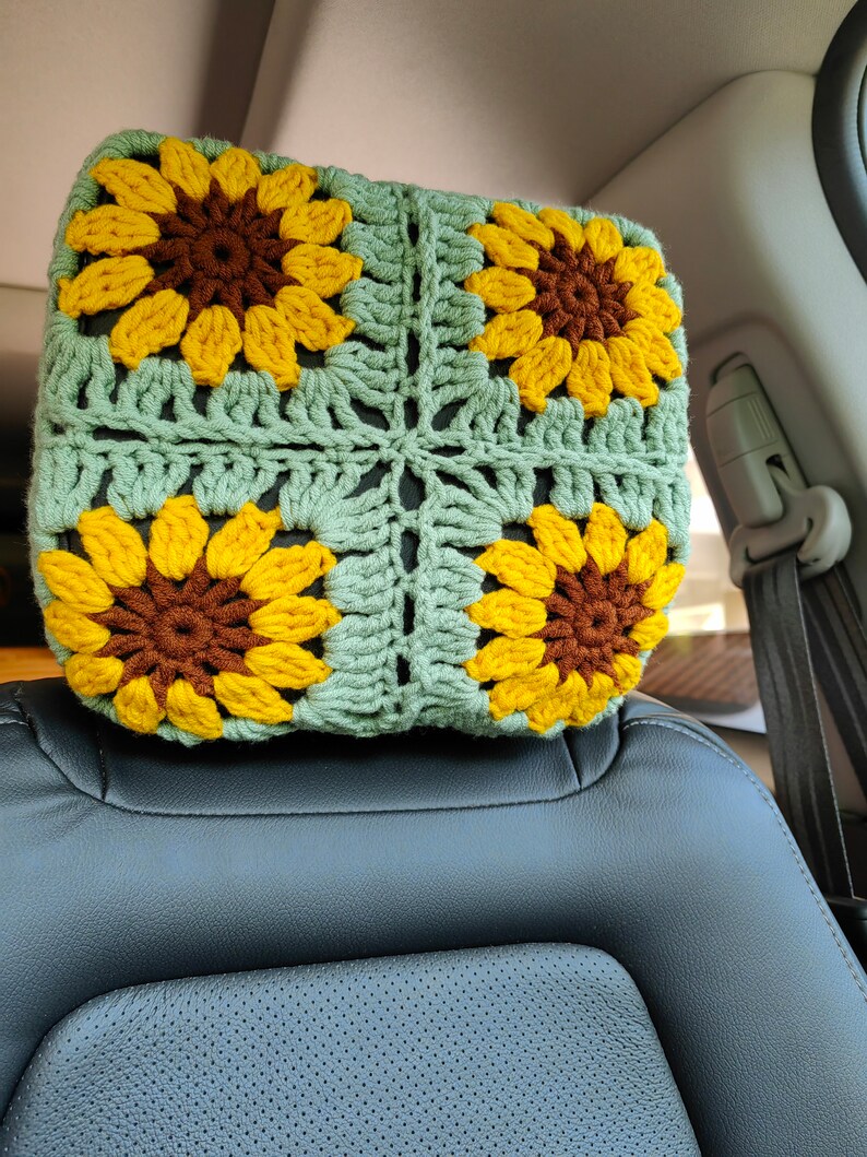 Puede incluir: Un reposacabezas de crochet con fondo verde y cuatro girasoles amarillos con centros marrones. El reposacabezas est&aacute; en un coche.