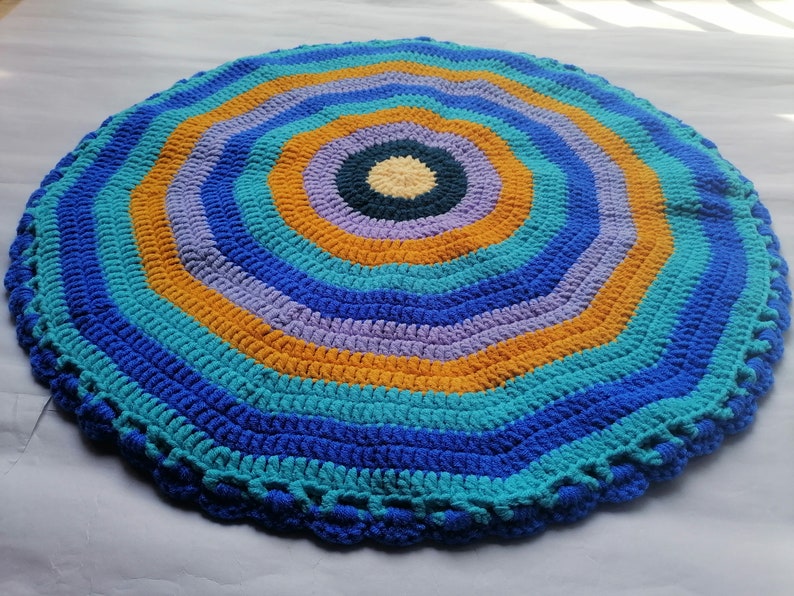 May include: A round crocheted rug with a multi-colored concentric circle pattern. The rug is made with blue, teal, orange, and purple yarn.