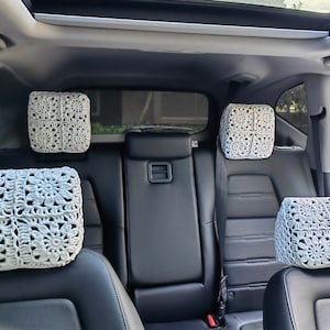 May include: White crocheted headrest covers for car seats. The covers are made with a square pattern and are placed on the headrests of the back seats of a car.
