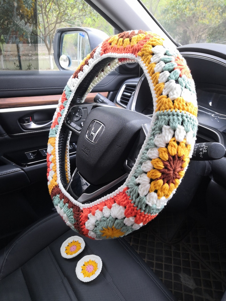 May include: A colorful crocheted steering wheel cover with a floral pattern in shades of orange, yellow, brown, white, and green. The cover is made of yarn and has a textured surface. Two matching crocheted coasters are also included.
