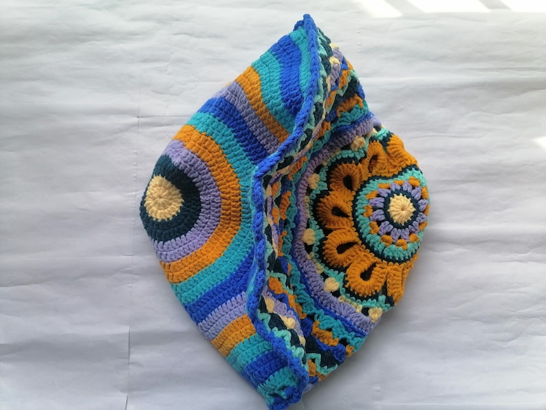 May include: A crocheted hat with a colorful circular pattern in shades of blue, purple, yellow, and orange. The hat is folded in half, revealing the intricate design.