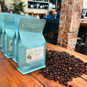 May include: Several light blue bags of coffee beans are arranged on a wooden surface, with a pile of roasted coffee beans to the right. The bags have a label that reads "Holiday Blend" and "Deans Beans".