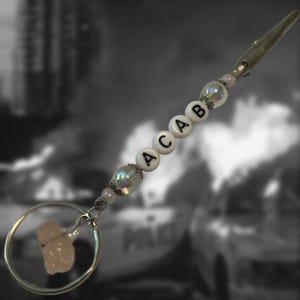 May include: A silver-toned clip with a beaded strand spelling out "ACAB." The strand includes white letter beads, iridescent beads, and pink beads. A small, irregular-shaped, light pink stone hangs from a silver keyring.
