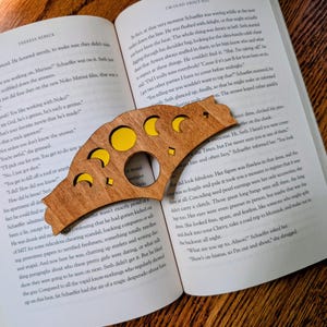 May include: A wooden bookmark with a crescent moon and phase of the moon design. The bookmark is placed on an open book with text. The phases of the moon are cut out and filled with yellow.