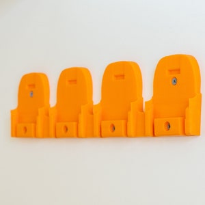 Puede incluir: Cuatro soportes de pared de color naranja brillante. Cada soporte tiene una parte superior curvada, una peque&ntilde;a abertura rectangular en la parte inferior y un tornillo para el montaje. Los soportes est&aacute;n espaciados uniformemente y fijados a una pared blanca.