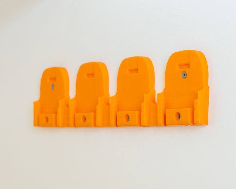 Puede incluir: Cuatro soportes de pared de color naranja brillante. Cada soporte tiene una parte superior curvada, una peque&ntilde;a abertura rectangular en la parte inferior y un tornillo para el montaje. Los soportes est&aacute;n espaciados uniformemente y fijados a una pared blanca.