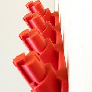 K&ouml;nnte beinhalten: Ein roter, an der Wand montierter Batteriehalter mit mehreren Schlitzen. Der Halter besteht aus Kunststoff und ist f&uuml;r mehrere Batterien ausgelegt. Der Halter ist an einer wei&szlig;en Wand befestigt.