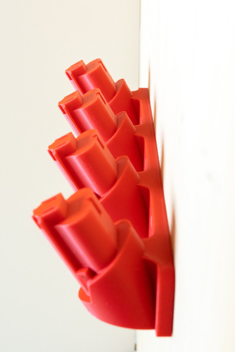 K&ouml;nnte beinhalten: Ein roter, an der Wand montierter Batteriehalter mit mehreren Schlitzen. Der Halter besteht aus Kunststoff und ist f&uuml;r mehrere Batterien ausgelegt. Der Halter ist an einer wei&szlig;en Wand befestigt.