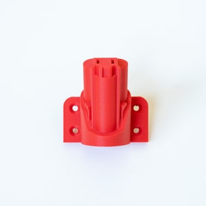 K&ouml;nnte beinhalten: Ein leuchtend roter, 3D-gedruckter Werkzeugbatteriehalter. Der Halter hat ein zylindrisches Design mit einer rechteckigen Basis mit Befestigungsl&ouml;chern. Das Objekt dient zur Aufnahme einer Elektrowerkzeugbatterie. Der Halter ist aus Kunststoff.