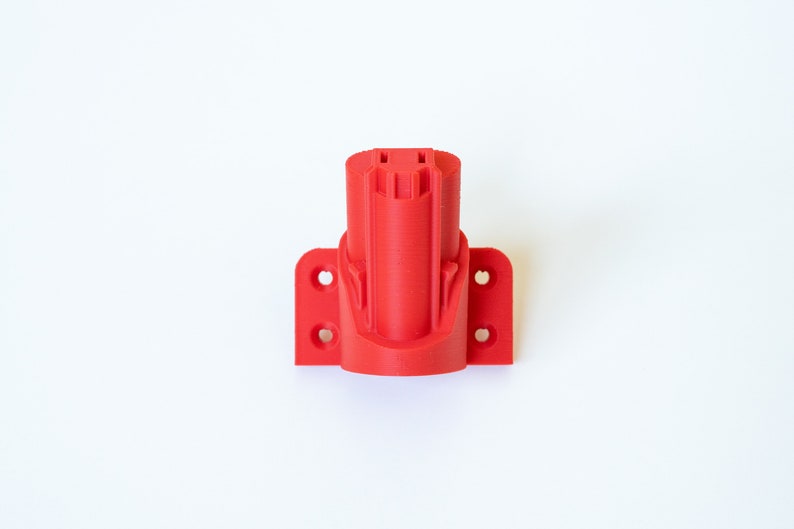 K&ouml;nnte beinhalten: Ein leuchtend roter, 3D-gedruckter Werkzeugbatteriehalter. Der Halter hat ein zylindrisches Design mit einer rechteckigen Basis mit Befestigungsl&ouml;chern. Das Objekt dient zur Aufnahme einer Elektrowerkzeugbatterie. Der Halter ist aus Kunststoff.