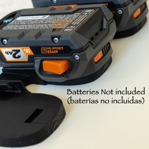 Puede incluir: Bater&iacute;as Hyper Lithium Ion de 18V negras y naranjas con una capacidad de 2Ah. Los paquetes est&aacute;n sujetos a un soporte negro. El texto "Batteries Not included" es visible.