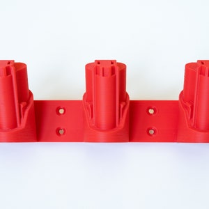 K&ouml;nnte beinhalten: Ein roter 3-fach Batteriehalter f&uuml;r Elektrowerkzeuge. Der Halter besteht aus Kunststoff und hat drei zylindrische Schlitze zur Aufnahme von Batterien. Der Halter ist f&uuml;r die Montage an einer Wand oder einer anderen Oberfl&auml;che konzipiert.