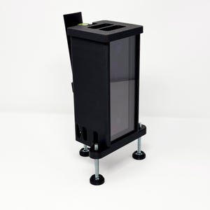 May include: A black rectangular device with a clear front panel and adjustable feet. The device has a top section with two recessed slots and a small level indicator. The feet are attached with bolts.