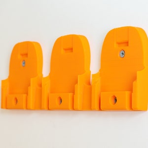 Puede incluir: Tres soportes de pared de color naranja brillante, cada uno con una forma &uacute;nica y un peque&ntilde;o agujero en la parte inferior. Los soportes se fijan a la pared con tornillos. El dise&ntilde;o parece estar impreso en 3D.