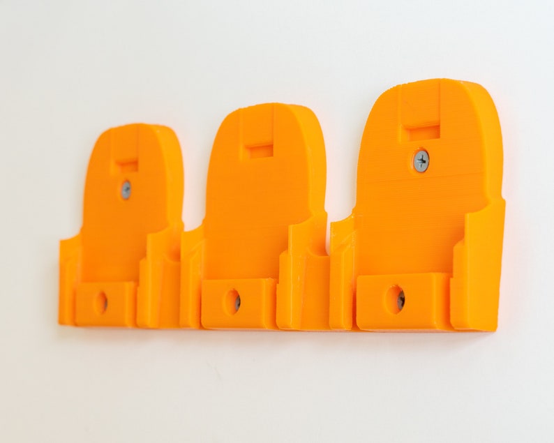 Puede incluir: Tres soportes de pared de color naranja brillante, cada uno con una forma &uacute;nica y un peque&ntilde;o agujero en la parte inferior. Los soportes se fijan a la pared con tornillos. El dise&ntilde;o parece estar impreso en 3D.