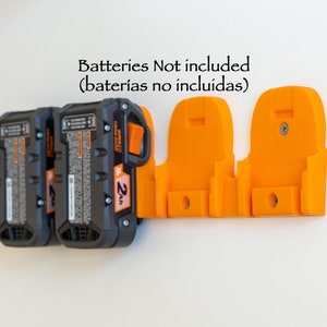 Puede incluir: Un soporte de bater&iacute;a naranja impreso en 3D montado en una pared blanca, que sostiene tres bater&iacute;as de herramientas el&eacute;ctricas negras y grises. El texto "Batteries Not included" es visible.