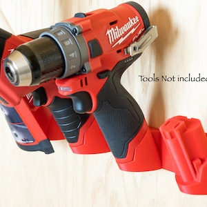 K&ouml;nnte beinhalten: Rote und schwarze Werkzeugaufbewahrung an einer hellen Holzoberfl&auml;che. Der Halter h&auml;lt eine Bohrmaschine und eine S&auml;bels&auml;ge. Der Text "Tools Not included" ist sichtbar.