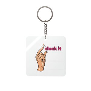 Puede incluir: Un llavero cuadrado blanco con un llavero y una cadena plateados. El llavero presenta una mano de dibujos animados que señala las palabras "clock it" en rosa. El diseño es simple y moderno.