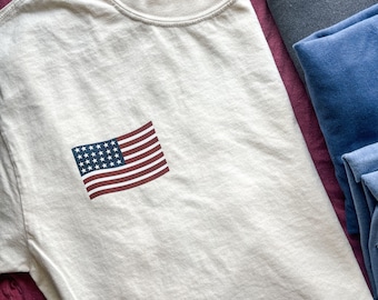 May include: A white t-shirt with a small American flag printed on the front. The shirt has a crew neck and short sleeves. The tag on the shirt reads "Comfort Colors" and "L-G-G".