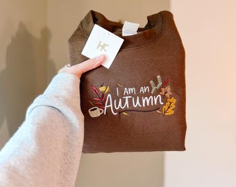 May include: A brown sweatshirt with the embroidered phrase "I am an Autumn" in white script, accompanied by fall-themed leaf and coffee cup designs. A white tag with "HC" is attached.