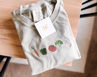 May include: A light green t-shirt with embroidered vegetables, including a carrot, bell pepper, tomato, broccoli, and garlic. The shirt is folded and has a tag that says "Comfort Colors" and "L-G-G".