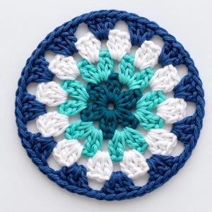 May include: A handmade, circular crochet doily in shades of blue, teal, and white. The center is a deep teal, surrounded by teal and white petals, and framed by a navy blue border. The doily is a decorative item.
