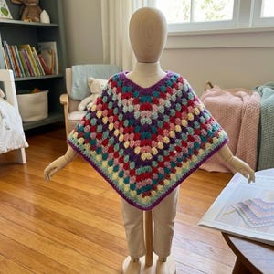 Può includere: Un poncho colorato all'uncinetto fatto a mano con un motivo a granny square. Il poncho, con scollo a V, è esposto su un manichino di legno. La tavolozza dei colori comprende viola, rosso, verde acqua, giallo e crema, con un bordo viola.
