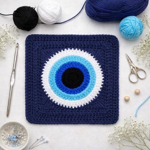 Könnte beinhalten: Ein marineblau gehäkeltes Quadrat mit einem weißen, hellblauen und dunkelblauen Evil-Eye-Design. Das Quadrat ist von Garn, einem Häkelhaken, einer Schere und Stecknadeln umgeben. Die Garnfarben sind Marineblau, Weiß, Schwarz und Hellblau.