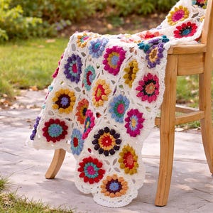 May include: A handmade, off-white crochet blanket draped over a wooden chair. The blanket features colorful floral motifs in shades of red, pink, yellow, blue, and purple. The blanket's design is a classic granny square pattern.