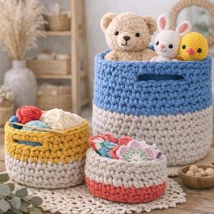 May include: A set of three handmade crochet baskets in varying sizes and colors. The largest basket is blue and white, holding a teddy bear, a white bunny, and a yellow chick. The other baskets are yellow and white, and coral and white, respectively.