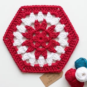 Può includere: Un sottobicchiere esagonale all'uncinetto rosso e bianco con un motivo floreale. Il sottobicchiere fatto a mano presenta un bordo rosso e dettagli bianchi. Un'etichetta recita "HAND CROCHETED". Sono visibili anche diversi gomitoli di filato rosso, bianco e verde acqua. Misura circa 15 cm.