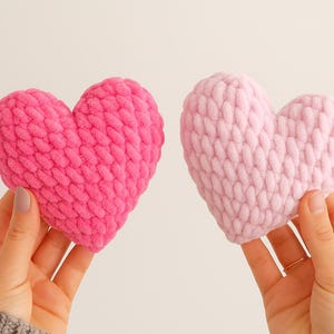 Può includere: Due cuscini fatti a mano a forma di cuore, uno fucsia vibrante e l'altro rosa pastello. Entrambi hanno una trama a maglia grossa e sono tenuti in mano. Lo sfondo è neutro. Un affascinante e accogliente oggetto artigianale.