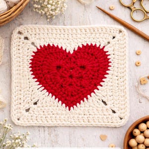Può includere: Un quadrato all'uncinetto bianco sporco con un vivace cuore rosso al centro. Il cuore è circondato da un bordo dello stesso filato bianco sporco. È probabilmente un oggetto fatto a mano, forse un sottobicchiere o un elemento decorativo.