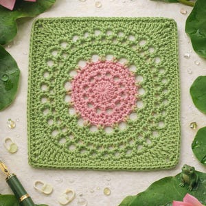 Puede incluir: Una pieza cuadrada de ganchillo hecha a mano con un diseño floral rosa central rodeado por un borde verde. La pieza está colocada entre hojas de nenúfar, un bolígrafo y una pequeña figura de rana, lo que sugiere un tema inspirado en la naturaleza.