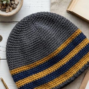 Puede incluir: Gorro de ganchillo gris hecho a mano con una banda horizontal de rayas azul marino y doradas. El gorro está hecho de hilo suave y es adecuado para el clima frío. Un accesorio elegante para cualquier armario.