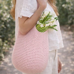 K&ouml;nnte beinhalten: Eine hellrosa geh&auml;kelte Tragetasche gef&uuml;llt mit wei&szlig;en Blumen. Die Tasche hat eine abgerundete Form und eine strukturierte Oberfl&auml;che. Die Blumen haben gr&uuml;ne Stiele und wei&szlig;e Bl&uuml;tenbl&auml;tter. Die Tasche wird von einer Person gehalten, die ein wei&szlig;es Hemd tr&auml;gt.