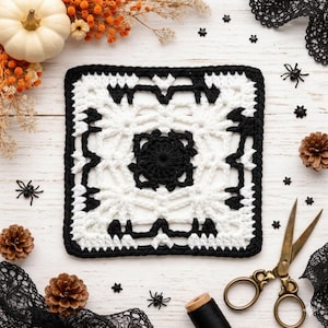 May include: A square, white and black crochet doily with a spiderweb design. The doily is surrounded by Halloween-themed decorations, including miniature spiders, pinecones, and a white pumpkin. A pair of gold scissors and black thread are also present.