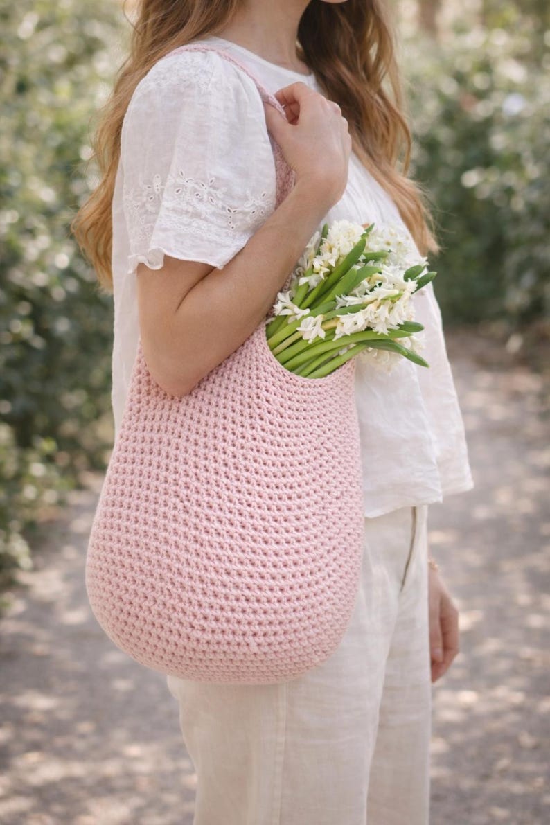 K&ouml;nnte beinhalten: Eine hellrosa geh&auml;kelte Tragetasche gef&uuml;llt mit wei&szlig;en Blumen. Die Tasche hat eine abgerundete Form und eine strukturierte Oberfl&auml;che. Die Blumen haben gr&uuml;ne Stiele und werden in der Tasche gehalten. Die Tasche wird von einer Person gehalten, die ein wei&szlig;es Hemd tr&auml;gt.