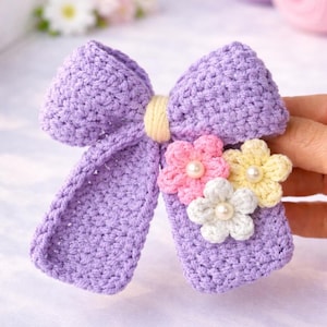 May include: A lilac-coloured, crocheted bow with a yellow band in the centre, adorned with three small, crocheted flowers in pink, white, and yellow, each with a pearl. Ideal for hair accessories or craft projects.