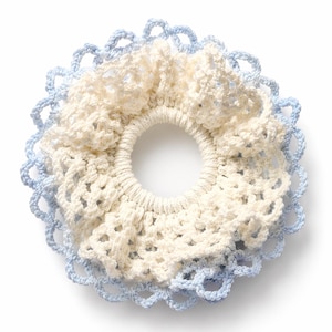 May include: A circular hair scrunchie featuring a central elastic band covered in cream-colored crochet fabric with a ruffled texture, surrounded by an outer border of light blue crochet lace with a repeating scalloped pattern throughout.