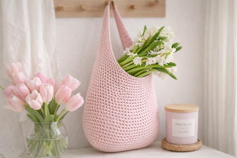 K&ouml;nnte beinhalten: Eine hellrosa geh&auml;kelte Tragetasche mit wei&szlig;en Blumen. Links steht eine Vase mit rosa Tulpen. Eine Kerze mit Holzdeckel und dem Text "Peony Blush" steht rechts auf einem gewebten Untersetzer.