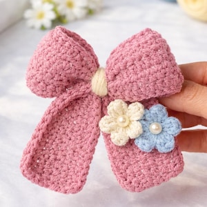 Può includere: Un fiocco rosa all'uncinetto fatto a mano con dettagli color crema. Due piccoli fiori all'uncinetto, uno color crema e uno azzurro, sono attaccati, ciascuno con un centro di perla. L'accessorio è per capelli.