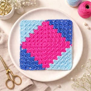May include: A square, handmade crochet piece in shades of pink, blue, and light blue, displayed on a white plate. The crochet pattern features a geometric design. Additional elements include yarn spools and small scissors.