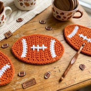 May include: Handmade orange crochet football coasters with white stitching details. The coasters are arranged on a wooden surface with small wooden football-shaped embellishments and a crochet hook. The scene includes mugs with fall-themed designs.