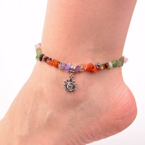 May include: Anklet with a strand of multi-coloured, irregularly shaped gemstones. A silver sun charm hangs from the centre. Gemstones include green, orange, purple, and brown. The anklet is worn on an ankle.